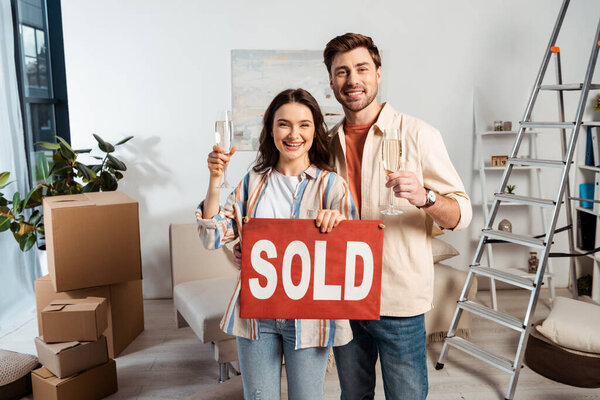 Smiling couple with glasses of champagne holding nameplate with sold lettering during moving in living room