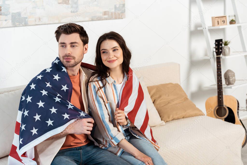 Young couple wrapped in american flag sitting on couch at home 