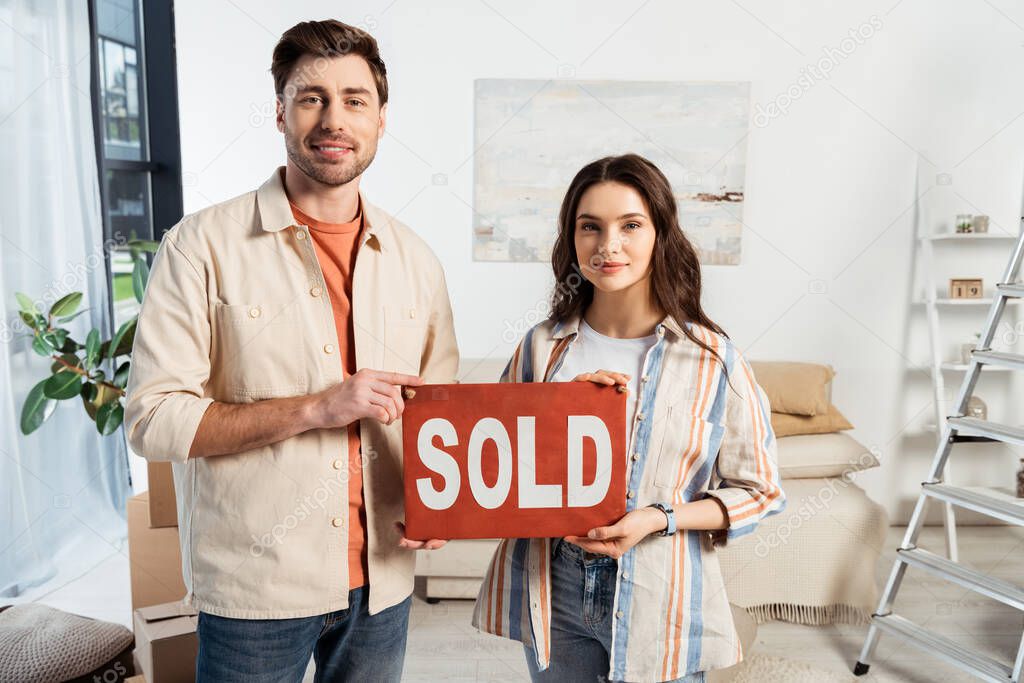 Smiling man holding nameplate with sold lettering near girlfriend in living room