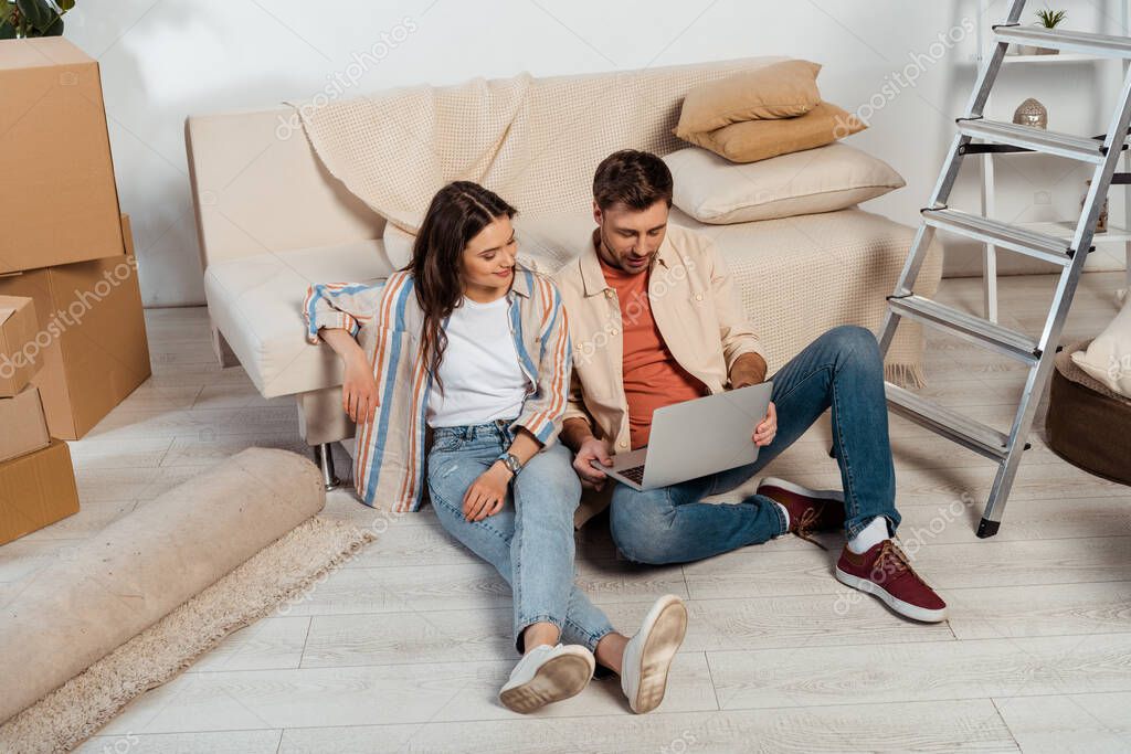 Smiling woman sitting near boyfriend using laptop in new house