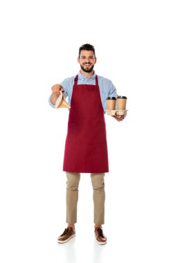 Handsome positive waiter holding paper bag and disposable cups on white background clipart