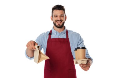 Positive waiter holding coffee to go and paper bag while looking at camera isolated on white clipart