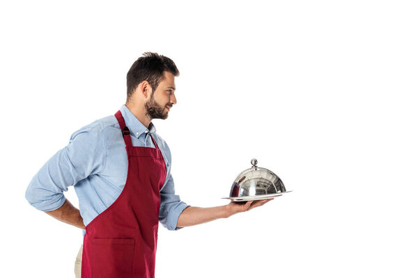 Красивый официант в фартуке с подносом и металлической крышкой, изолированной на белом
