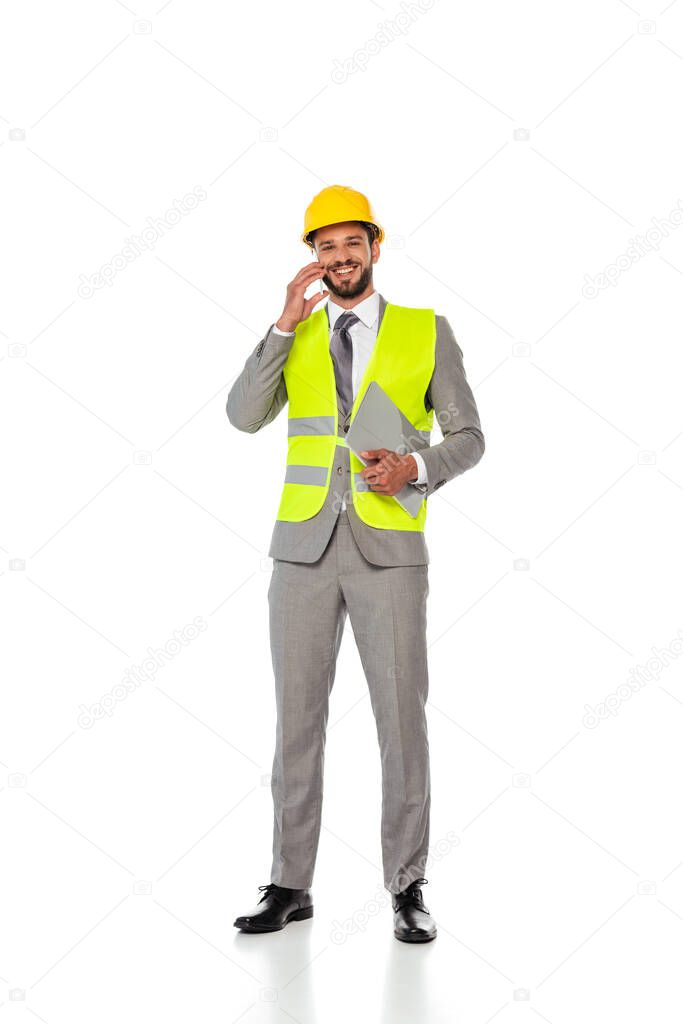 Smiling engineer in suit talking on smartphone and holding laptop on white background