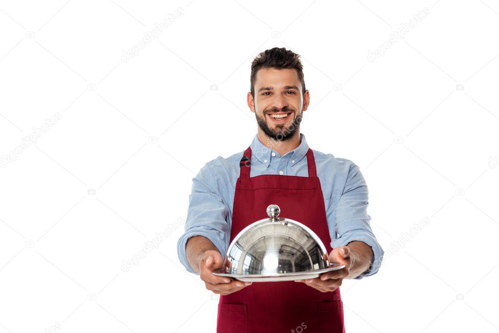 Smiling waiter in apron showing metal tray and dish cover isolated on white