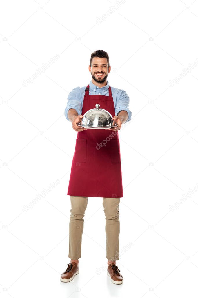 Handsome waiter smiling at camera and showing metal tray and dish cover on white background