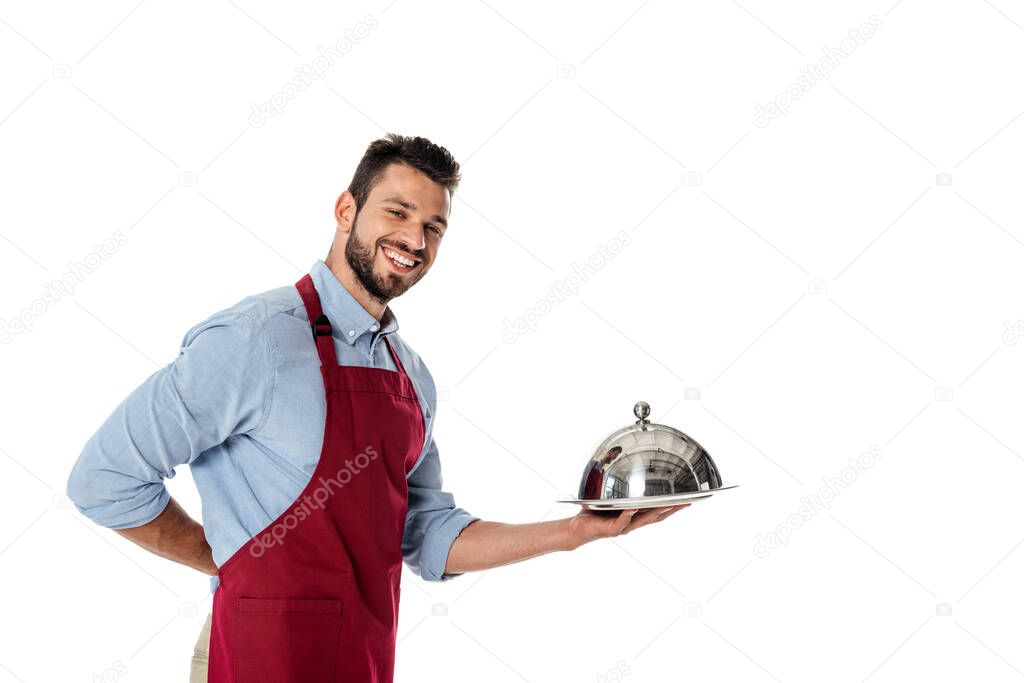 Waiter in apron holding metal dish cover and tray while smiling at camera isolated on white