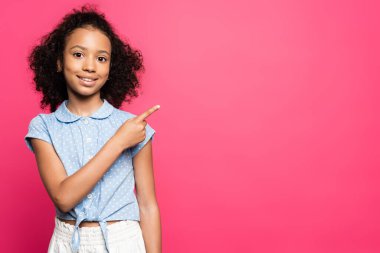smiling cute curly african american kid pointing with finger aside isolated on pink clipart