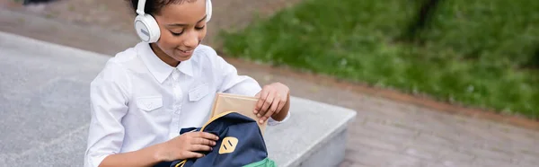 Happy African American Schoolgirl Headphones Putting Book Backpack Panoramic Shot — Stock Photo, Image
