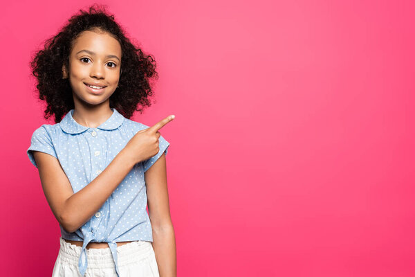 smiling cute curly african american kid pointing with finger aside isolated on pink