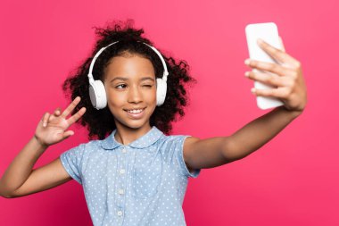 Gülümseyen kıvırcık saçlı, kulaklıklı Amerikalı çocuğun zafer işareti gösterip pembe renkteki akıllı telefondan selfie çektiği seçici odak noktası.