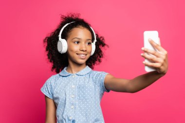 Gülümseyen kıvırcık saçlı, kulaklıklı Amerikalı çocuğun pembe renkli akıllı telefondan selfie çekmesi.