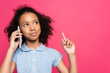 Gülümseyen kıvırcık Afrikalı Amerikalı çocuk akıllı telefondan konuşuyor ve pembe üzerine izole edilmiş bir fikir jesti gösteriyor.