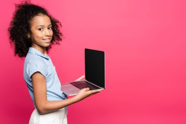 Gülümseyen kıvırcık kıvırcık saçlı Amerikalı çocuk elinde pembe ekranlı dizüstü bilgisayar tutuyor.