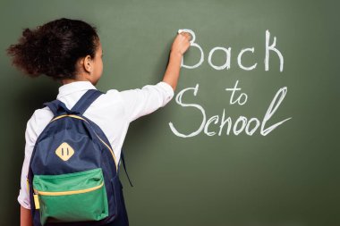 back view of african american schoolgirl with backpack writing back to school inscription on green chalkboard clipart