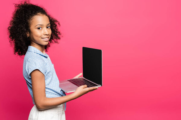 smiling cute curly african american kid holding laptop with blank screen isolated on pink