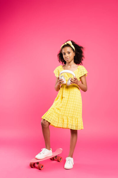 full length view of smiling curly african american child in yellow outfit on penny board with headphones on pink background