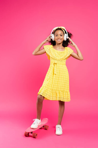 full length view of smiling curly african american child in yellow outfit and headphones on penny board on pink background