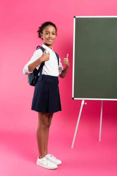 Sonriente Afroamericana Colegiala Con Mochila Cerca Pizarra Vacía Mostrando Pulgares — Foto de Stock