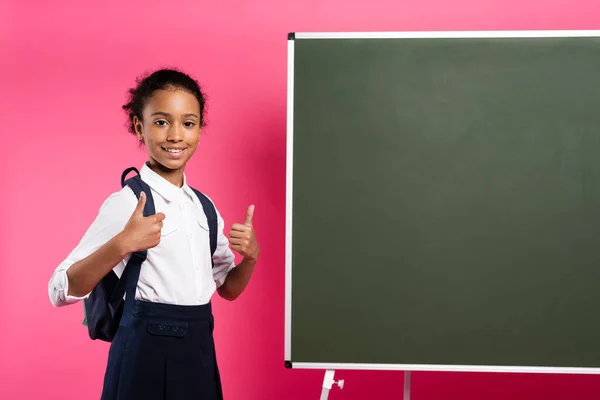 笑顔アフリカ系アメリカ人の女子学生ピンクの背景に親指を示す空の黒板の近くにリュックを持つ — ストック写真