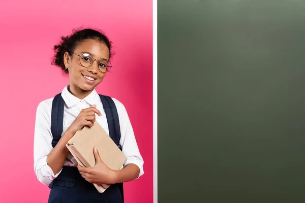 Усміхнена Афроамериканська Школярка Книгою Біля Порожньої Дошки Рожевому Фоні — стокове фото