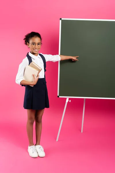 smiling african american schoolgirl with books pointing at empty chalkboard on pink background