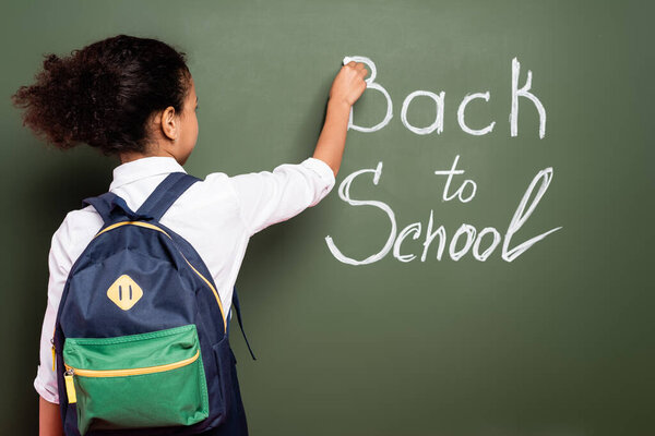 back view of african american schoolgirl with backpack writing back to school inscription on green chalkboard