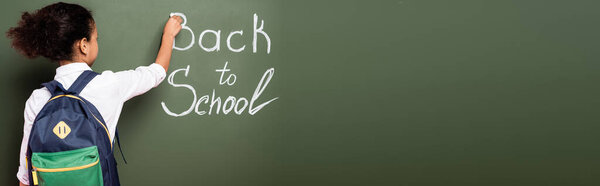 back view of african american schoolgirl with backpack writing back to school inscription on green chalkboard, panoramic shot