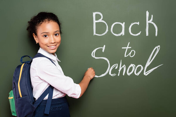 smiling african american schoolgirl with backpack writing back to school inscription on green chalkboard