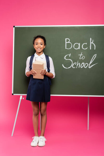 smiling african american schoolgirl with books near back to school inscription on green chalkboard on pink background