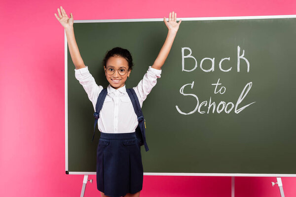happy african american schoolgirl in glasses with hands in air near back to school inscription on green chalkboard on pink background