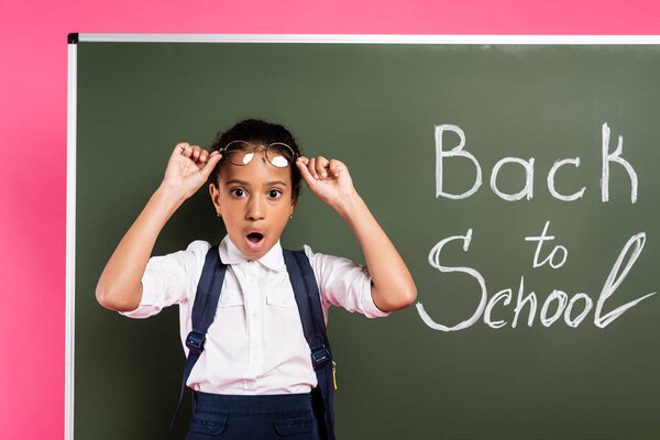 shocked  african american schoolgirl adjusting glasses near back to school inscription on green chalkboard on pink background