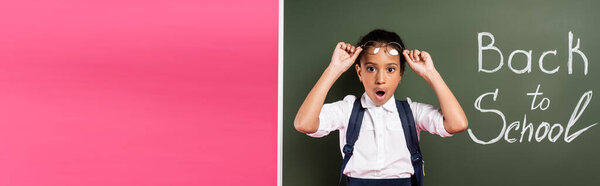 shocked african american schoolgirl adjusting glasses near back to school inscription on green chalkboard on pink background, panoramic shot