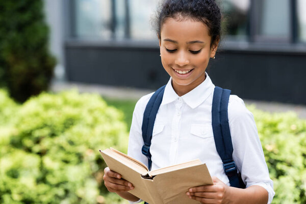 smiling african american schoolgirl reading book outdoors