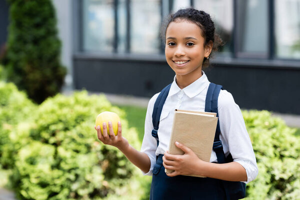 smiling african american schoolgirl with book and apple outdoors