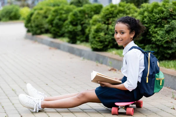 Zijaanzicht Van Glimlachend Afrikaans Amerikaans Schoolmeisje Lezen Boek Terwijl Zitten — Stockfoto