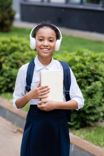 smiling african american schoolgirl in headphones with backpack and books outdoors