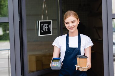 Gülümseyen garson elinde ödeme terminali ve kafenin kapısında açık harflerle tabelanın yanına gitmek için kahve tutuyor. 