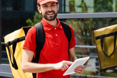 Sırt çantalı gülümseyen kurye şehir caddesinde dijital tablet kullanırken kameraya bakıyor 