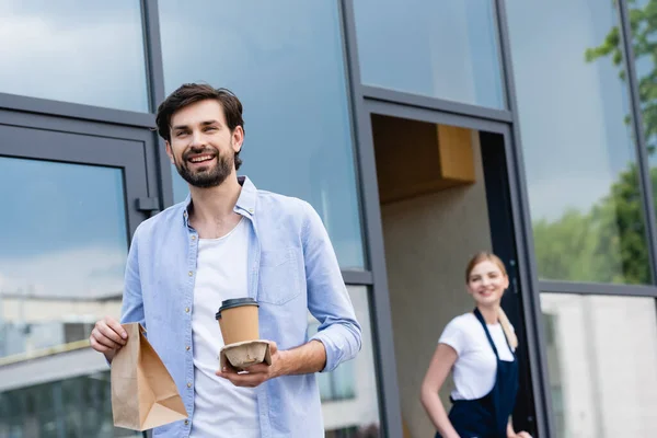 微笑的男人带着纸袋和咖啡 在城市街道的咖啡店附近与女服务员在一起的选择性焦点 — 图库照片