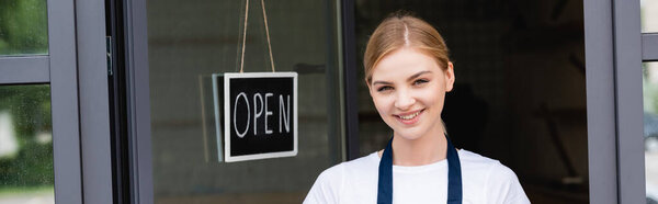 Panoramic shot of smiling waitress looking at camera near signboard with open lettering on door of cafe