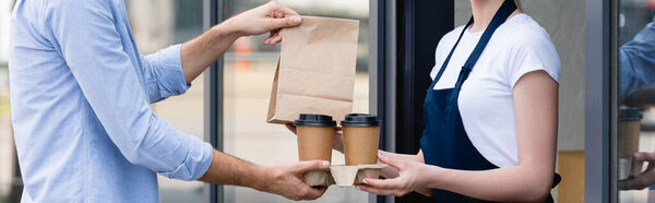 Panoramic crop of waitress giving disposable cups and paper bag to customer near cafe on urban street