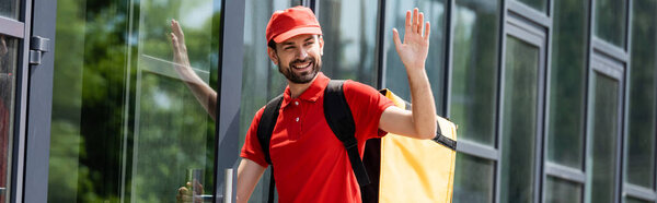 Panoramic shot of smiling delivery man waving hand near building on urban street 