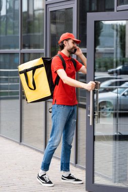 Smiling delivery man talking on smartphone and opening door of building on urban street 