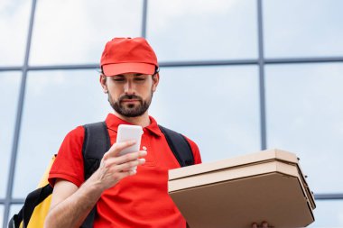 Pizza kutularını tutan kuryenin düşük açılı görüntüsü ve şehir caddesinde akıllı telefon kullanımı 