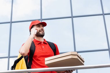 Sokak ortasında pizza kutularını tutarken akıllı telefondan gülümseyen kuryenin düşük açılı görüntüsü 