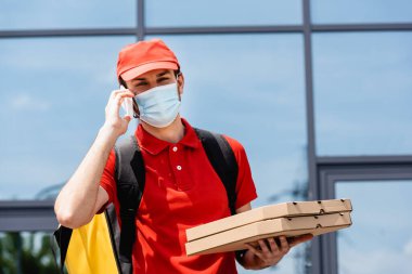 Tıbbi maskeli kurye akıllı telefondan konuşuyor ve şehir caddesinde pizza kutuları tutuyor. 