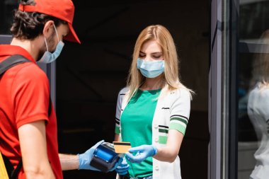 Tıbbi maskeli kadının seçici odak noktası, şehir caddesinde kurye ödeme terminalinin yanında kredi kartıyla ödeme yapması. 