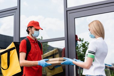 Tıbbi maskeli ve lateks eldivenli kurye, şehir caddesindeki binanın yakınındaki bir kadına pizza kutusu veriyor. 