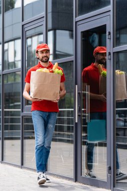 Smiling delivery man holding shopping bag with fresh vegetables while walking on urban street  clipart